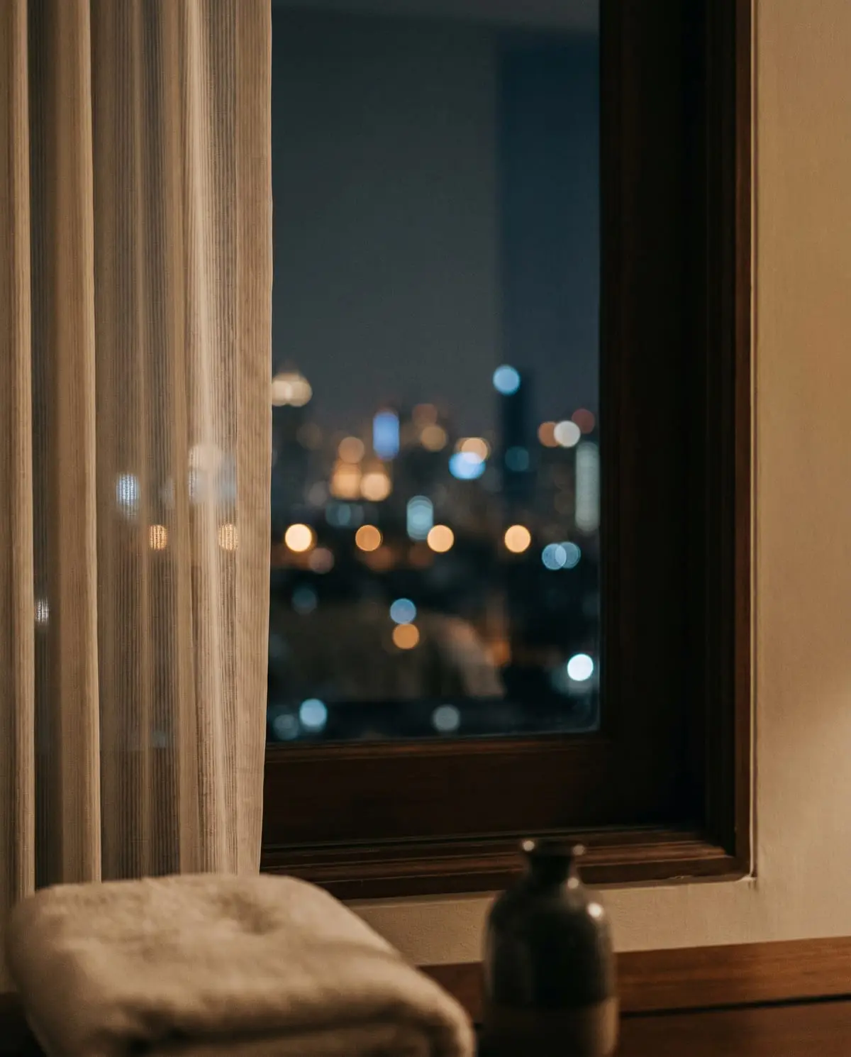 Bangkok's skyline glowing at night, seen through a sheer hotel curtain with a ceramic vessel on the sill