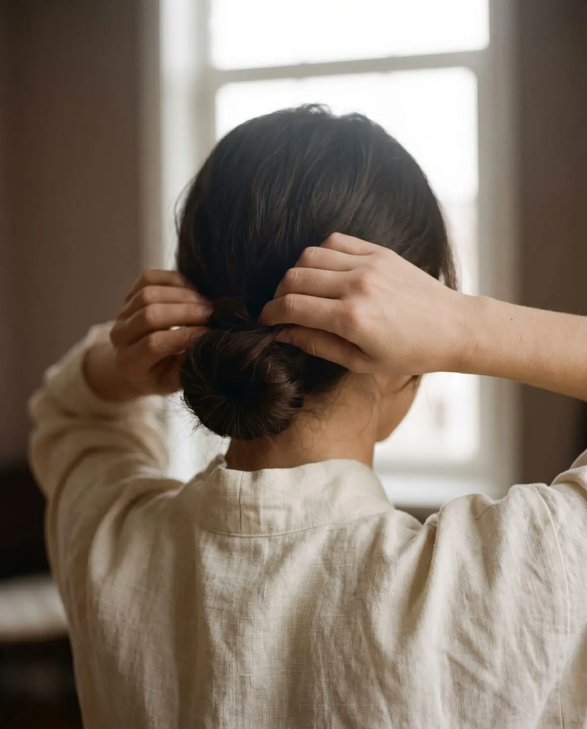 A therapist tying her hair into a low bun before a treatment, seen from behind in a linen shirt