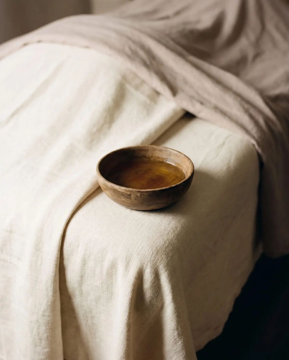 A wooden bowl of warm massage oil resting on a linen-draped treatment surface